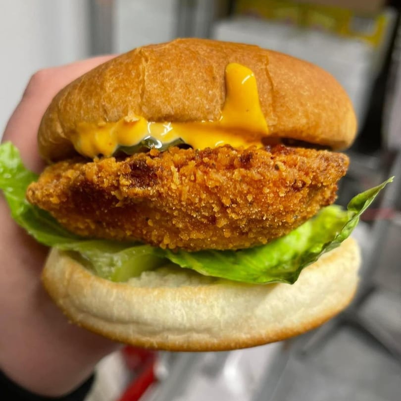 Hand holding a crispy fried chicken sandwich with lettuce, pickles, and cheese sauce on a bun.
