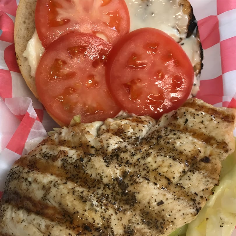 Grilled chicken sandwich with sliced tomatoes and mayonnaise on a bun, served in a red checkered paper.
